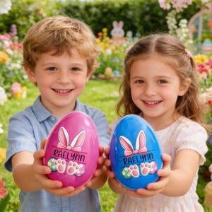 May include: Two children holding large, egg-shaped Easter decorations. One egg is pink, the other blue, both with bunny ears, a bow, the name "Raelynn", and colorful paw prints. The background features a garden with flowers and Easter decorations.