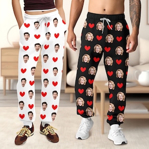 May include: Two pairs of sweatpants, one white and one black, each featuring a repeating pattern of red hearts and personalized faces. The white pants have a drawstring and are paired with brown sneakers. The black pants are worn with white sneakers.