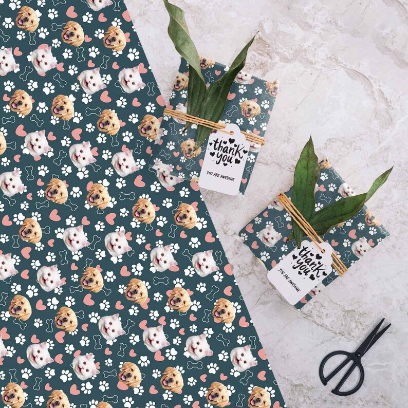 May include: A sheet of wrapping paper with a repeating pattern of dog and cat faces, pink hearts, white paw prints, and white bones on a dark teal background. The text "Thank You You Are Awesome" is on a white tag.
