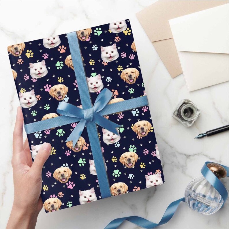 May include: A blue gift box with a blue ribbon wrapped around it. The box is covered in a dark blue wrapping paper with a pattern of paw prints and cat and dog faces. The paw prints are in various pastel colors.