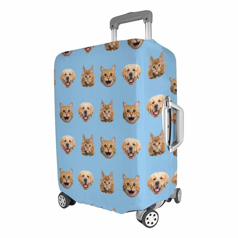 May include: A light blue suitcase cover with a repeating pattern of smiling golden retriever and cat faces. The suitcase has a silver handle and wheels, designed for travel and luggage protection.