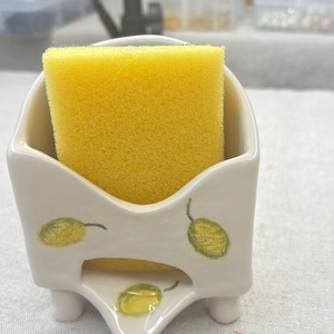 Puede incluir: Porta esponjas de cerámica blanca con diseño de limón. El porta esponjas tiene tres patas y una esponja amarilla en su interior.