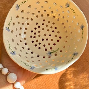 Puede incluir: Cuenco de cerámica color crema con numerosas perforaciones circulares. El cuenco está adornado con delicados acentos florales azules y tallos verdes. El diseño pintado a mano añade un toque único a esta pieza decorativa. Ideal para frutas o como objeto decorativo.
