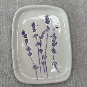 Puede incluir: Un pequeño plato de cerámica blanca con forma cuadrada y bordes redondeados. El plato presenta un diseño de flores de lavanda en tonos morados.