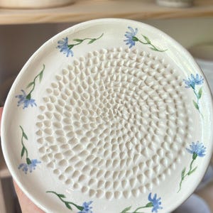 以下が含まれることがあります： 中央にすりおろし用の突起がある白い円形の陶器製おろし皿。皿の縁には、緑の茎と葉を持つ小さな青いヤグルマギクが手描きで円状に描かれており、すりおろし面は細かい突起が密集したテクスチャーになっています。