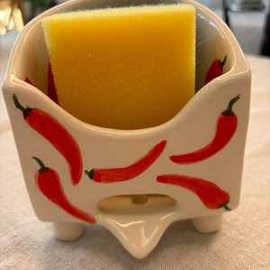 Peut inclure: Porte-éponge en céramique blanche avec des motifs de piments rouges. Le porte-éponge a une petite ouverture au fond pour le drainage. Une éponge jaune est placée à l'intérieur du porte-éponge.