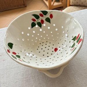 May include: White ceramic colander with a pattern of green leaves and red berries. The colander has many small holes for draining and a handle on each side. A kitchenware item.