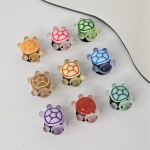 Könnte beinhalten: Eine Sammlung farbenfroher Schildkröten-Anhänger. Jeder Anhänger hat einen andersfarbigen Panzer, darunter Orange, Rosa, Grün, Blau, Braun, Gelb, Rot, Lila und Türkis. Die Anhänger haben eine glänzende Oberfläche und sind auf einer weißen Oberfläche angeordnet.