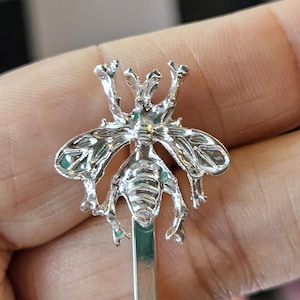 Puede incluir: Una pinza para el pelo plateada con forma de abeja. La abeja tiene alas y segmentos corporales detallados. La pinza es de estilo horquilla, ideal para sujetar el cabello. El diseño es intrincado y detallado.