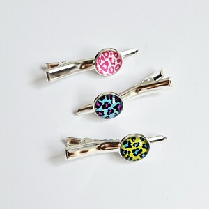 Puede incluir: Tres pinzas para el pelo plateadas con diseños redondos y coloridos de estampado de leopardo. Una pinza presenta un patrón rosa y blanco, otra tiene un diseño azul y morado, y la tercera un patrón amarillo y negro. Accesorios para el cabello.