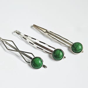 Op de afbeelding: Drie zilveren haarclips met groene, marmerachtige, ronde accenten. De clips hebben verschillende ontwerpen: één is ruitvormig en de andere twee zijn recht. Op een witte achtergrond.