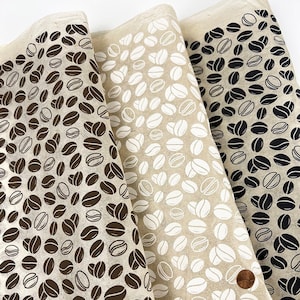 May include: Three pieces of fabric with a coffee bean pattern. The fabric on the left has brown coffee beans on a beige background. The fabric in the middle has white coffee beans on a beige background. The fabric on the right has black coffee beans on a beige background.