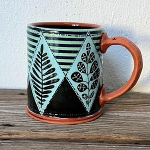 K&ouml;nnte beinhalten: Eine Keramiktasse mit einem geometrischen Muster in Schwarz, Wei&szlig; und Blau mit stilisierten Bl&auml;ttern. Die Tasse hat einen braunen Henkel und einen braunen Rand.