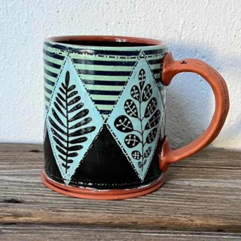 K&ouml;nnte beinhalten: Eine Keramiktasse mit einem geometrischen Muster in Schwarz, Wei&szlig; und Blau mit stilisierten Bl&auml;ttern. Die Tasse hat einen braunen Henkel und einen braunen Rand.