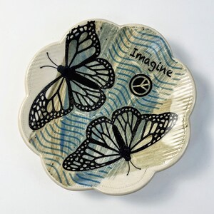 May include: A white ceramic dish with a scalloped edge, featuring two black butterfly silhouettes and the word "Imagine" with a peace sign. The background is a blue and white striped pattern.