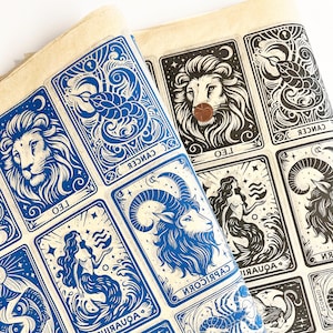 Op de afbeelding: Twee vellen papier met illustraties van dierenriemtekens. Eén vel is blauw en wit, de andere zwart en wit. Elke kaart toont een ander dierenriemteken, zoals een leeuw, een zeemeermin en een ram. De kaarten zijn rechthoekig met afgeronde hoeken.