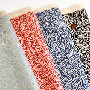 Op de afbeelding: Vier rollen stof met een herhalend abstract patroon van gebogen lijnen en halve cirkels. De kleuren van de stof zijn lichtblauw, rood, donkerblauw en zwart, allemaal gedrukt op een natuurlijke beige canvas basis. Een koperen munt is zichtbaar.