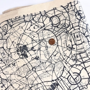 Puede incluir: Tela blanquecina con un diseño abstracto negro con líneas, círculos y formas. Se coloca una moneda de cobre en la tela para la escala. La tela tiene un aspecto texturizado y un borde en capas.