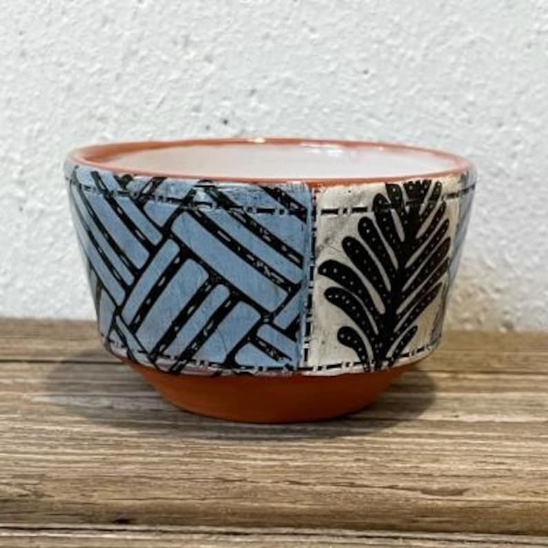 K&ouml;nnte beinhalten: Eine kleine, keramische Schale mit einem blauen und wei&szlig;en geometrischen Muster und einem schwarzen und wei&szlig;en Blatt-Muster. Die Schale ist aus Terrakotta und hat eine leicht abgerundete Form.