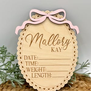 Puede incluir: Letrero de anuncio de nacimiento de madera con el nombre "Mallory Kay" en escritura cursiva. El letrero tiene un borde festoneado y un lazo rosa en la parte superior. Incluye espacios para registrar la fecha, hora, peso y longitud del beb&eacute;.