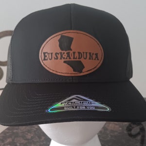 May include: Black trucker hat with a brown leather patch that says "EUSKALDUNA" and a California state outline. The hat has a black mesh back and a "Pacific Headwear Built For You" label.