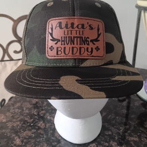 May include: Camouflage trucker hat with a brown leather patch that says "Aita's Little Hunting Buddy" in black lettering.