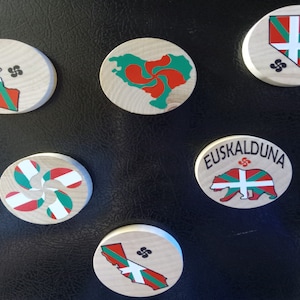 May include: Six round wooden magnets with different designs featuring the Basque flag, a bear, and a stylized Basque cross. The magnets are on a black surface.