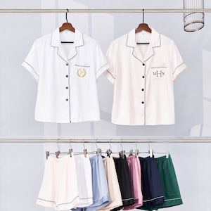 Puede incluir: Dos tops de pijama de satén blanco y rosa con ribetes negros y cierres de botón. La parte superior blanca tiene una "P" bordada en oro con una corona de laurel. La parte superior rosa tiene una "MHN" bordada en negro. Ocho pares de pantalones cortos de pijama de satén en varios colores, incluyendo rosa, blanco, azul, beige, negro, morado, azul marino y verde.