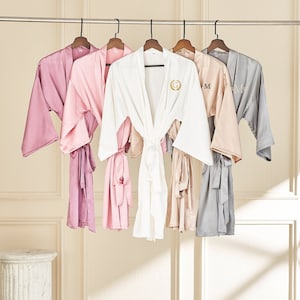Könnte beinhalten: Fünf Satin-Bademäntel hängen an einem Gestell. Die Bademäntel sind in verschiedenen Farben: rosa, weiß, hellrosa, beige und grau. Jeder Bademantel hat eine Satinschleife und ein Monogramm auf der Vorderseite.