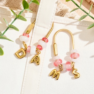 Può includere: Due chiusure metalliche dorate a forma di U con perline decorative in rosa traslucido e bianco opaco. Ogni chiusura è decorata con due ciondoli a forma di lettera in metallo dorato, che compongono rispettivamente le lettere D, A, M e S.