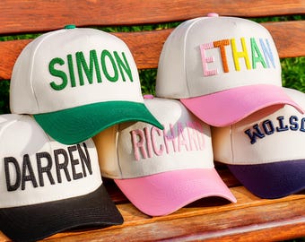 May include: A collection of personalized baseball caps with names embroidered on the front. The caps feature a white crown, colored brims, and names in various colors. The names include "SIMON", "ETHAN", "DARREN", "RICHARD", and "CUSTOM".