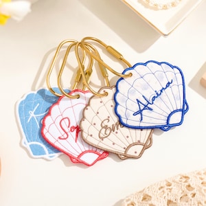 Peut inclure: Quatre porte-clés en forme de coquillage brodé avec des anneaux en métal doré. Chaque coquillage présente une couleur unique—bleu, rouge, beige et bleu foncé—et est personnalisé avec des noms brodés comme Alaina, Emma et Sophia.
