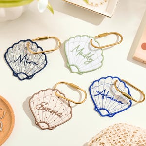 Op de afbeelding: Vier schelpvormige sleutelhangers met goudkleurige metalen ringen. Elke hanger heeft een geborduurde naam: Mira in donkerblauw, Maddy in lichtgroen, Emma in bruin en Alaina in blauw. De schelpen hebben diverse patronen zoals bloemen, ruiten en stippen.