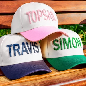Op de afbeelding: Drie baseballpetten met witte kronen en gekleurde kleppen worden getoond. Eén pet heeft een roze klep en het woord "TOPSAIL" geborduurd in roze. Een andere heeft een marineblauwe klep met "TRAVIS" in marineblauw. De derde heeft een groene klep met "SIMON" in groen.