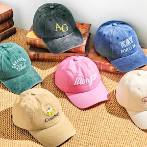 Könnte beinhalten: Eine Auswahl von sechs Baseballkappen in verschiedenen Farben, darunter Grün, Blau, Rosa und Beige. Jede Kappe ist mit gesticktem Text oder Initialen versehen, wie "Dreamers Club", "K&Y Forever" und "Morgan". Die Kappen liegen auf einer gewebten Oberfläche.