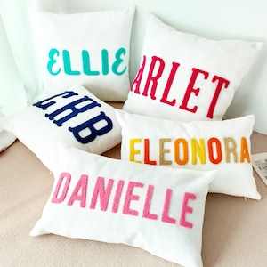 Puede incluir: Cojines blancos con nombres bordados en varios colores. Los nombres incluyen "ELLIE" en turquesa, "ARLET" en rosa, "CHB" en azul marino, "ELEONORA" en amarillo, naranja y beige, y "DANIELLE" en rosa.