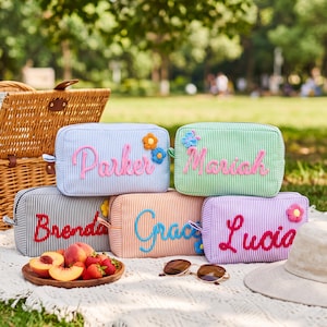 May include: Five rectangular zipped pouches in blue, green, black, orange, and purple pinstriped fabric. Each pouch features a personalised name in raised, colourful script lettering and small fabric flower appliqués attached to the front.