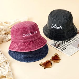 May include: Three bucket hats in dark grey, magenta, and navy blue with a washed denim texture. The grey hat features the embroidered white text "Bride" and the magenta hat features the embroidered white text "Custom Text". Heart-shaped sunglasses.