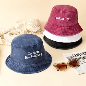 Może przedstawiać: Niebieski kapelusz typu bucket hat z białym haftowanym napisem Custom Embroidery, obok stosu trzech kapeluszy w kolorze bordowym, białym i czarnym oraz okularów przeciwsłonecznych w kształcie serc. Bordowy kapelusz ma biały haftowany napis Custom Text.