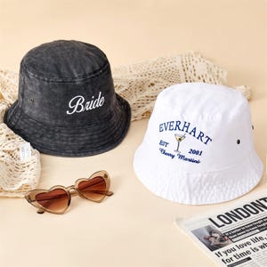 Puede incluir: Un sombrero de pescador negro con efecto lavado al ácido con la palabra Bride bordada en blanco, un sombrero blanco con el texto azul EVERHART EST 2001 Cherry Martini y un gráfico de copa de martini, y gafas de sol en forma de corazón.