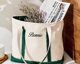 Könnte beinhalten: Eine cremefarbene Tragetasche aus Segeltuch mit dunkelgrünen Rändern und Griffen. Der Name Bonnie ist in dunkelgrüner Schreibschrift auf der Vorderseite eingestickt. Die Tasche enthält getrocknetes Grün und eine Zeitung mit dem Text London und If you love life.