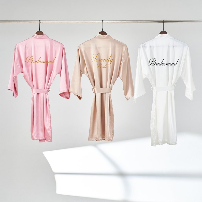 May include: Three satin robes hanging on hangers. The robe on the left is pink with the word "Bridemaid" embroidered in gold. The robe in the middle is beige with the words "Serenity Bride" embroidered in gold. The robe on the right is white with the word "Bridemaid" embroidered in black.