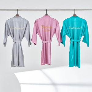 May include: Three satin robes hanging on hangers. The robe on the left is light gray with the word "Bridemaid" embroidered on the back. The robe in the middle is pink with the words "Victoria Bride" embroidered on the back. The robe on the right is turquoise with the word "Bridemaid" embroidered on the back.