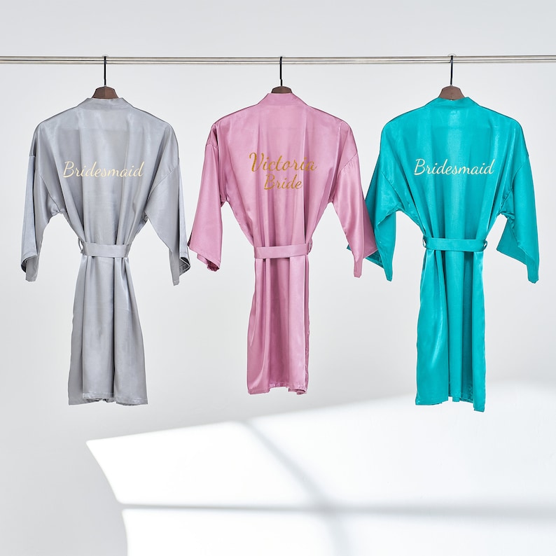 May include: Three satin robes hanging on hangers. The robe on the left is light gray with the word "Bridemaid" embroidered on the back. The robe in the middle is pink with the words "Victoria Bride" embroidered on the back. The robe on the right is turquoise with the word "Bridemaid" embroidered on the back.