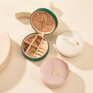 Puede incluir: Un estuche de viaje para joyas de terciopelo verde con cremallera dorada, abierto para revelar anillos y un collar. Se muestran dos estuches de joyería redondos adicionales, uno blanco con "Mumu" bordado y un corazón, y uno rosa con una "S" dorada.