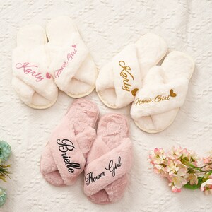 May include: Three pairs of fluffy slippers with crossover straps. The slippers are white, pink, and white. Each pair has the words "Flower Girl" embroidered on them. The white slippers have the name "Karly" embroidered on them. The pink slippers have the name "Brielle" embroidered on them.