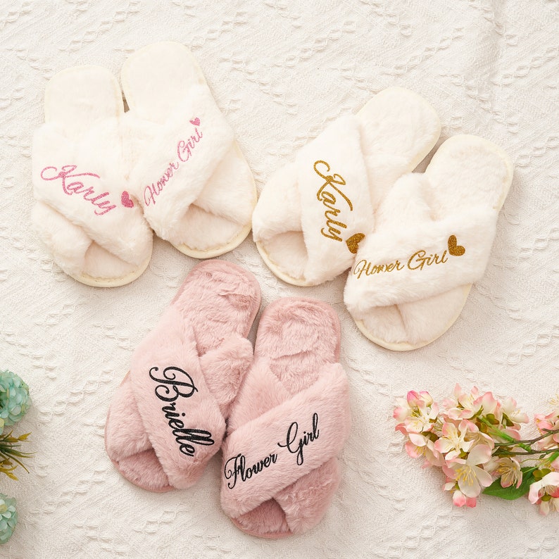 May include: Three pairs of fluffy slippers with crossover straps. The slippers are white, pink, and white. Each pair has the words "Flower Girl" embroidered on them. The white slippers have the name "Karly" embroidered on them. The pink slippers have the name "Brielle" embroidered on them.