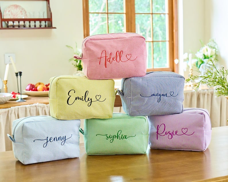 K&ouml;nnte beinhalten: Sechs personalisierte Kosmetiktaschen in verschiedenen Farben: Rosa, Gelb, Blau, Gr&uuml;n und Lila. Jede Tasche hat einen anderen Namen in Schreibschrift. Die Taschen haben ein gestreiftes Muster.