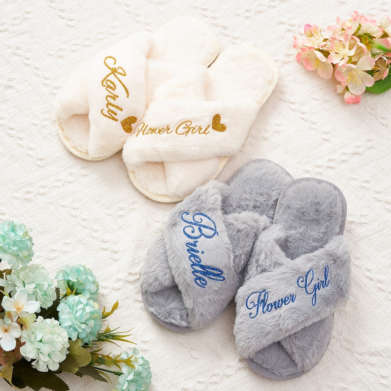 May include: Two pairs of fluffy slippers with crossover straps. One pair is white with gold glitter lettering that says "Karly Flower Girl". The other pair is grey with blue glitter lettering that says "Brielle Flower Girl".