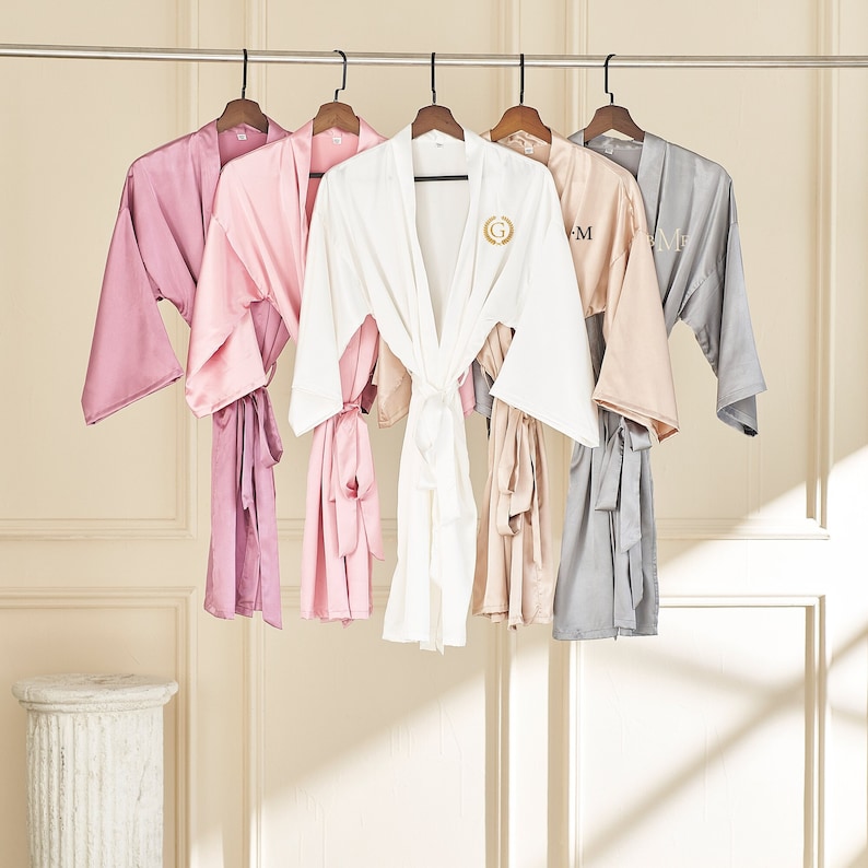 May include: Five satin robes hanging on a rack. The robes are in different colors: pink, white, light pink, beige, and gray. Each robe has a satin sash and a monogram on the front.